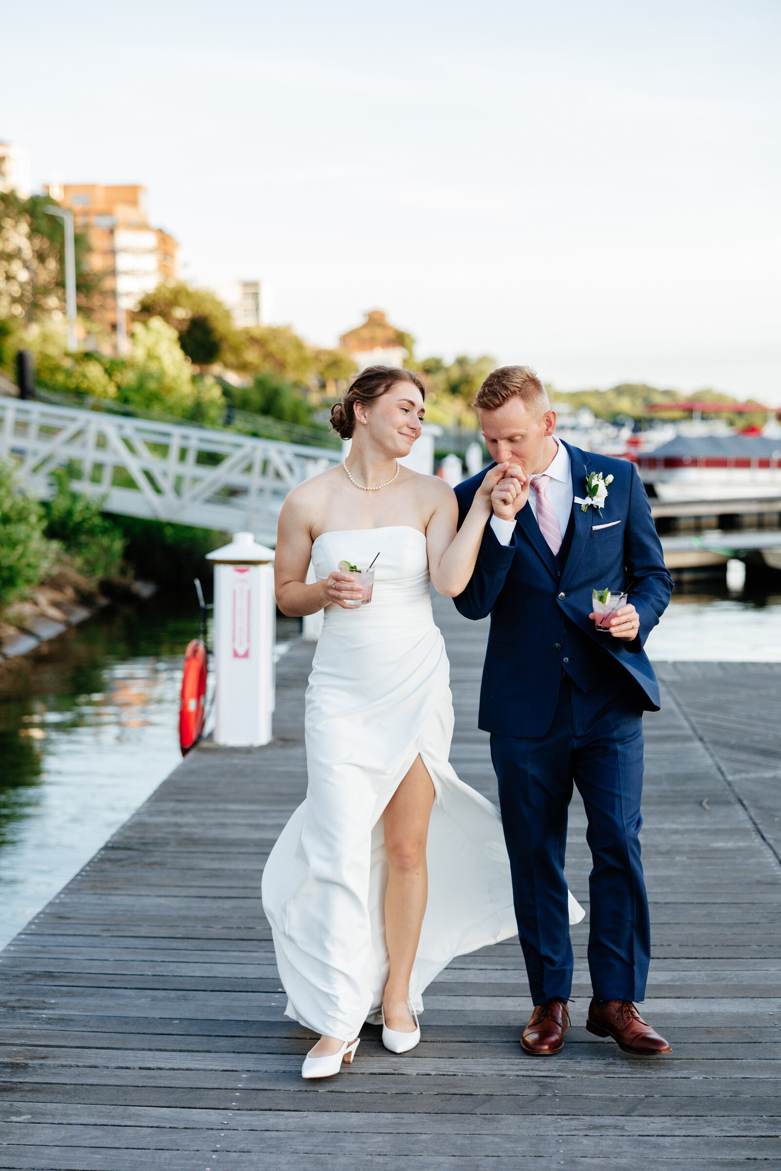 Groom kissing his bride’s hand as they walk along a marina dock