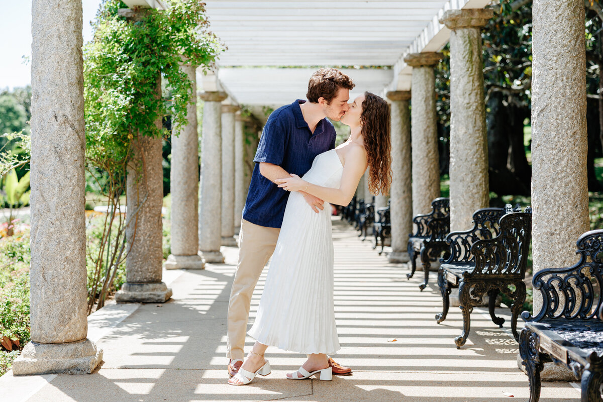 Engaged couple kissing beside white columns and a garden balustrade