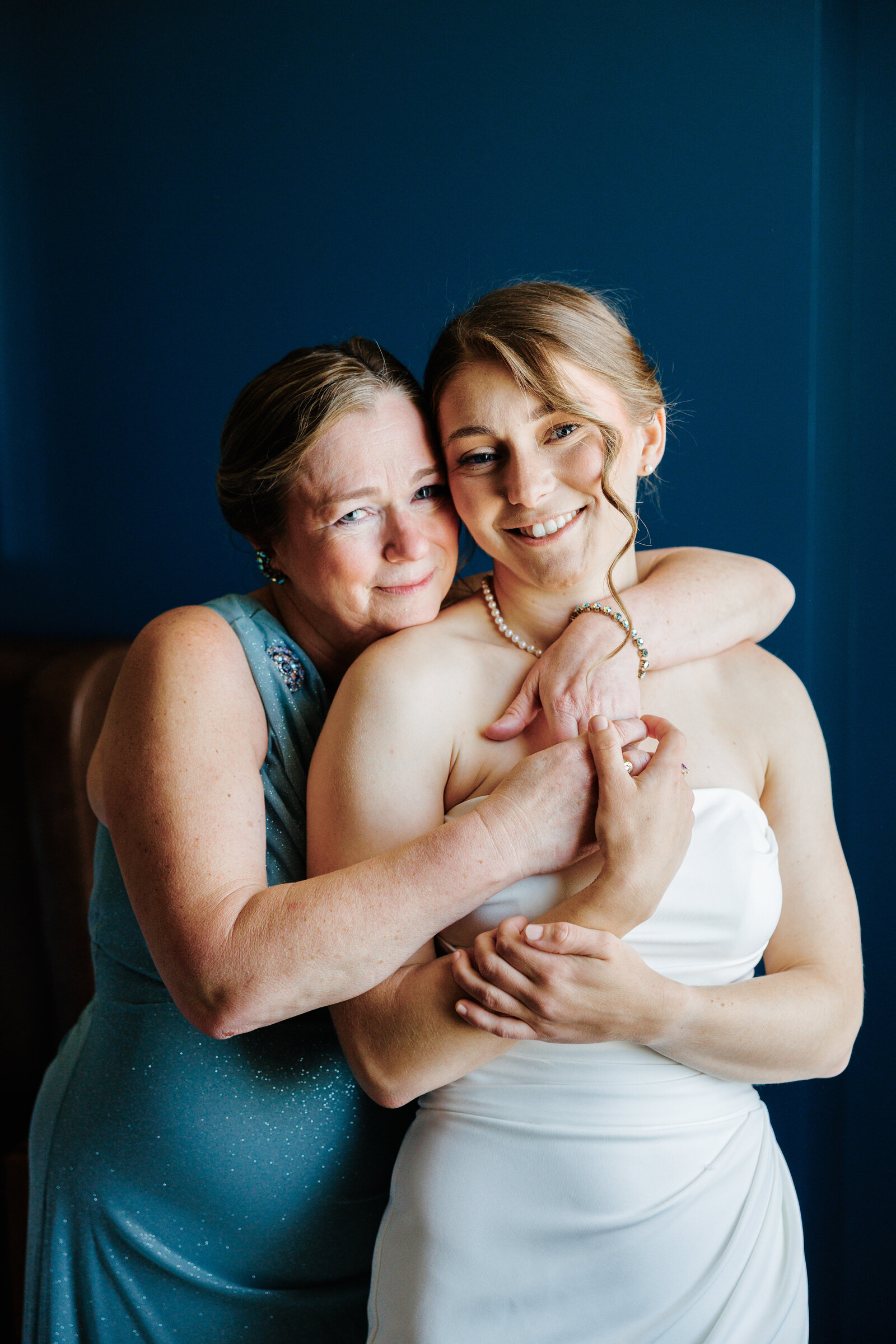 Mother hugging the bride from behind against a blue wall