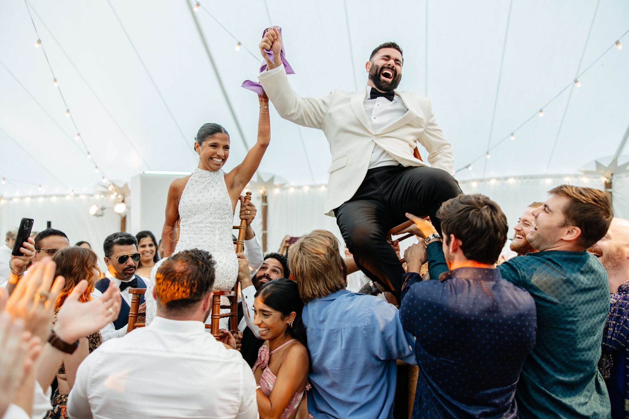Bride and groom lifted on chairs in a reception tent as guests cheer