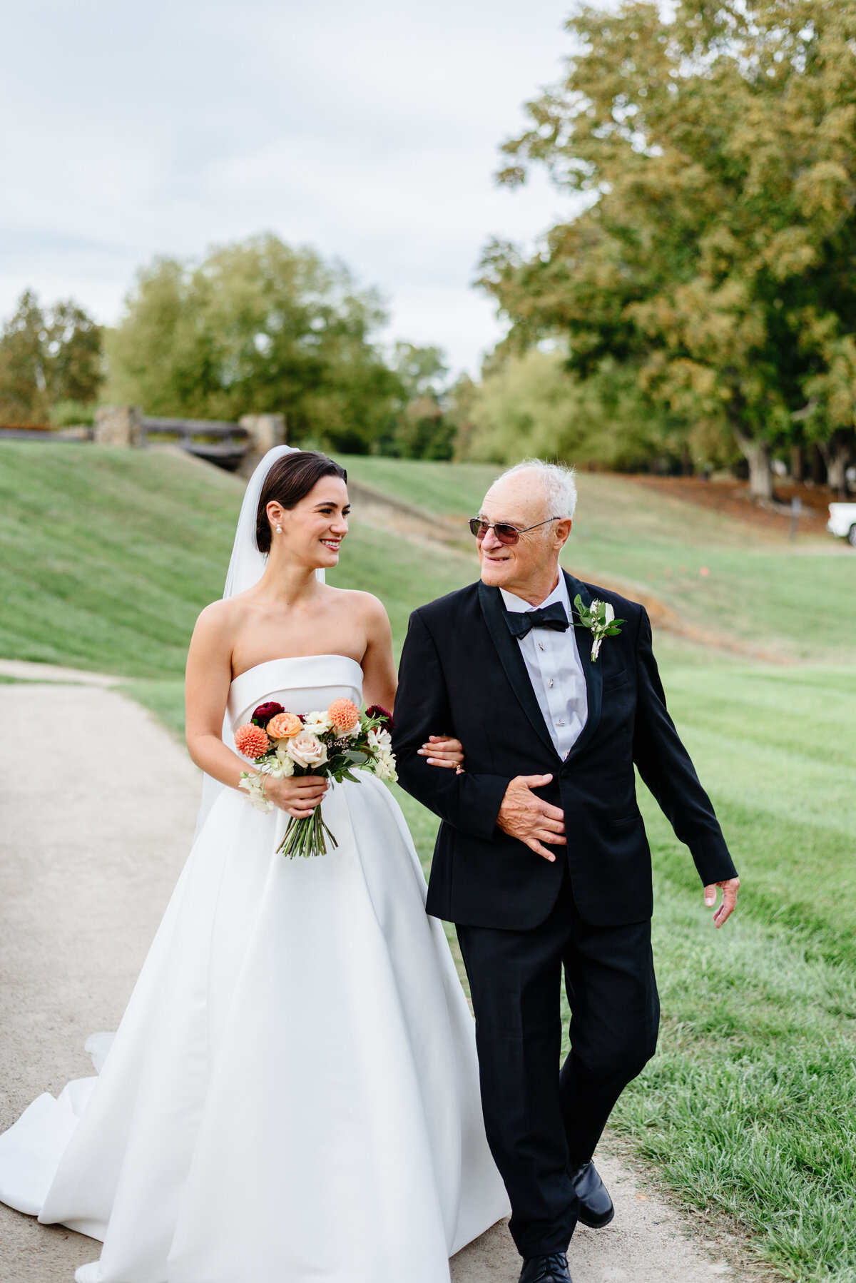 Bride walking arm in arm with an older man across a grassy ceremony lawn